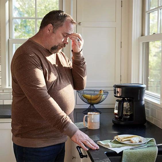 The image shows a man standing in a kitchen, holding his forehead in a gesture that may indicate stress, headache, or fatigue. 